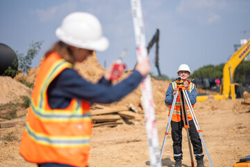 Construction surveyor or engineer working construction site blueprint and theodolite mounted tripod measuring angles and distances with heavy machinery excavator constructed hi-way road background.