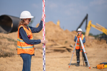 Construction surveyor or engineer working construction site blueprint and theodolite mounted tripod measuring angles and distances with heavy machinery excavator constructed hi-way road background.