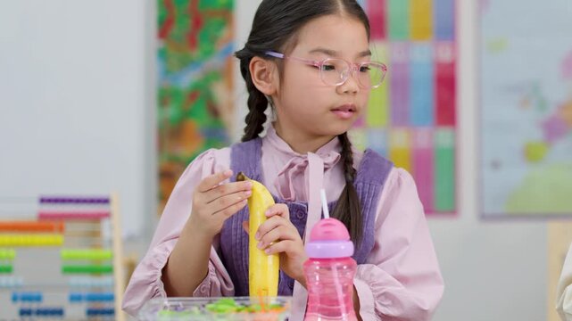Asian schoolgirl peeling banana during classroom snack time