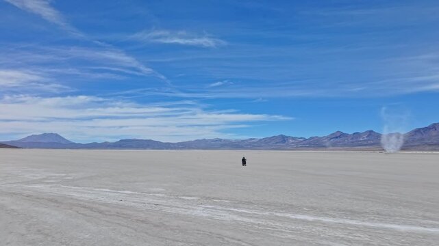 A massive salt tornado twists across the flats as two motorbike riders circle behind it, fearlessly riding straight toward the swirling storm in the remote Andes.