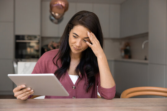 Woman sitting at table in modern kitchen, holding tablet and touching forehead with worried, disappointed expression due to slow internet connection, software crash, low battery, viruses or malware