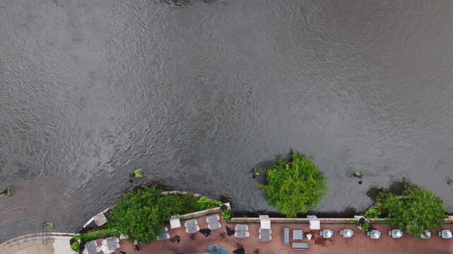 Aerial view of a swollen river flowing through a city after heavy rainfall. High water levels fill the river channel near walkways, showing flooding area and conditions