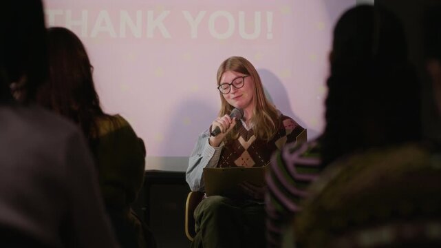 Medium shot of young woman holding microphone and speaking during presentation in front of audience, sharing ideas and insights in creative professional event setting