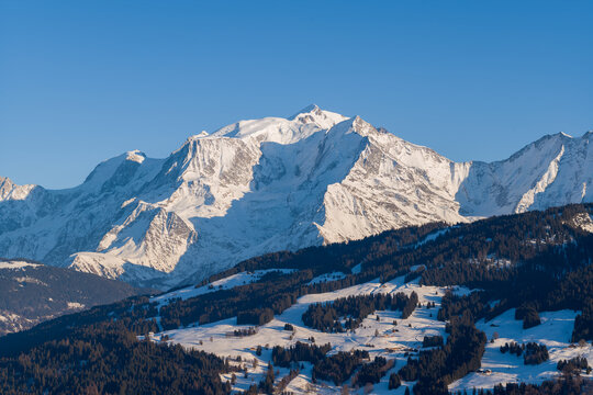 The towering snow-covered peaks of the Mont Blanc Massif rise above sunlit alpine foothills dotted with forests and scattered chalets under a clear blue sky.