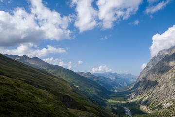 Naklejka premium Vast view of Val Ferret showing steep green hillsides, a winding river, and rugged peaks beneath a sky filled with scattered clouds and bright sunlight.