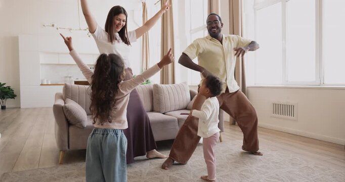 Cheerful interracial couple dancing, moving to music playfully with two biracial children in cozy living room, enjoying movement, laughter, and family bonding. Active leisure, happy parenthood, hobby