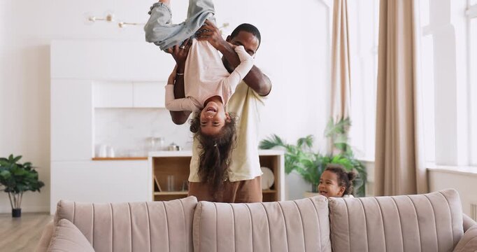 Happy Black father lifting his joyful biracial daughters upside down and put on sofa, laughing, playing together in modern living room, spend weekend at home, enjoy active games. Family time with kids