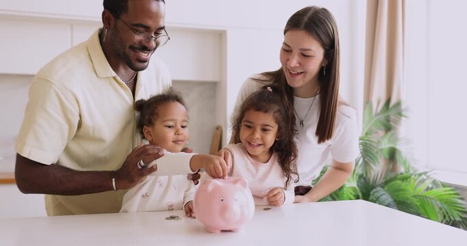 Interracial couple looking how two biracial little kids placing coins into piggybank, teaching to save money, early financial literacy, life habit, responsibility, standing together in kitchen at home