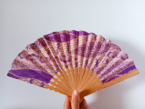 Close up of woman hand holding a purple and beige Indonesian traditional batik handheld folding fan or kipas tangan with a bamboo frame isolated on white background.