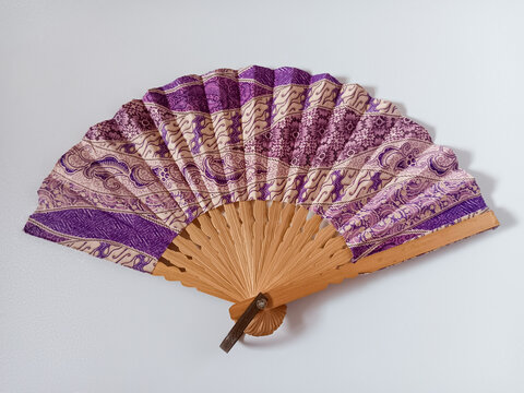 Close up view of a purple and beige Indonesian traditional batik handheld folding fan or kipas tangan with a bamboo frame isolated on white background.