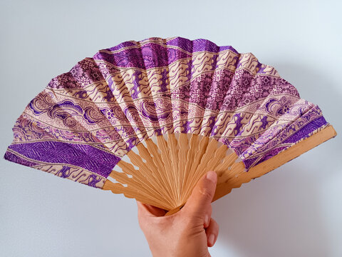 Close up of woman hand holding a purple and beige Indonesian traditional batik handheld folding fan or kipas tangan with a bamboo frame isolated on white background.