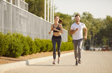 Fototapeta premium Sporty fit man and woman couple running and smiling together on paved path outdoors, happy exercising in shared active lifestyle, health and vitality positive energy in sunny day, fitness fun