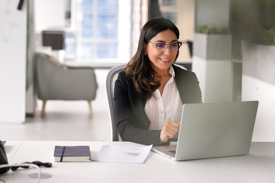 Smiling mature woman in glasses working on financial report, researching or websurfing information seated at desk with modern laptop in contemporary corporate office. Innovative AI business tech usage