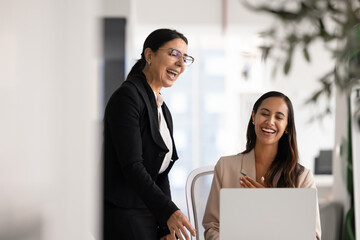 Fototapeta premium Mature 60s and young 30s women colleagues laughing in office while watching amusing content on laptop, take break during workday, giggling, enjoy fun and entertaining video in contemporary workspace