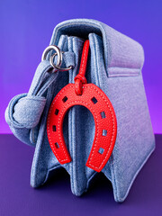 A close-up of a red leather bag charm in the shape of a horseshoe, placed on  blue bag