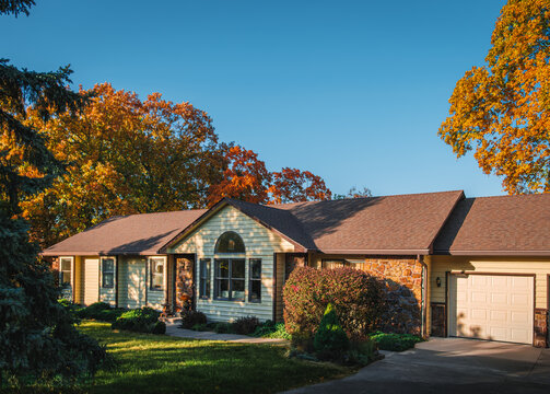 View of Midwestern suburban house before sunset in autumn; large colorful trees and clear sky behind house; long shadows on the lawn