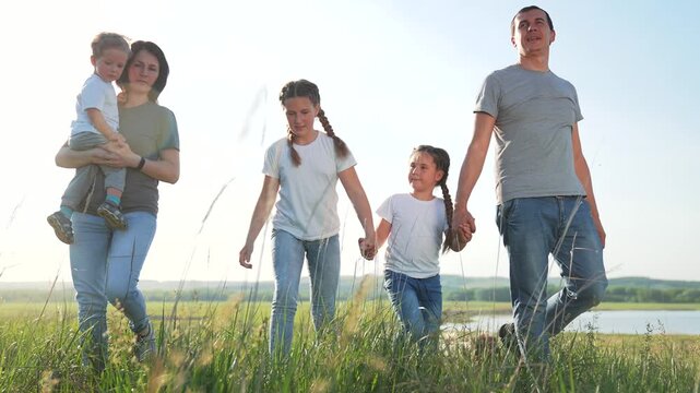 Family walk in meadow by lake with father and mother holding daughter and son while parent carries toddler child and grass sways under sky and distant hill reflects on calm water joyful outing