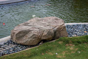 Large grey ornamental boulder by a pond with river pebbles and green grass. Serene outdoor landscaping feature showing a natural stone at the water's edge. © Ink N Propeller