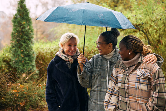 Smiling mature woman holding umbrella while spending leisure time with friends in outdoors rain