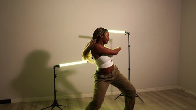Young energetic african woman with braids dancing modern afrodance. Joyful female performer moving to the rhythm in a studio with lights