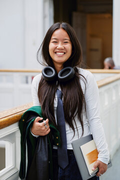 Portrait of smiling young woman with wireless headphones around her neck standing in university corridor