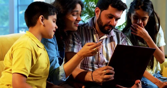 Indian young family watching old photo album at home revisiting cherished memories together while sitting on sofa indoors inside a modern lavish living room enjoying laughter bonding nostalgia
