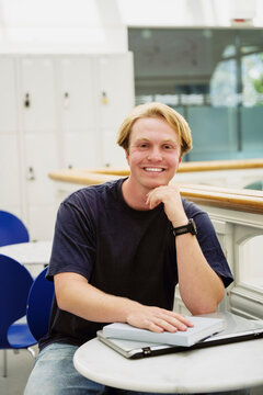 Portrait of smiling male university student sitting at table with study materials in campus