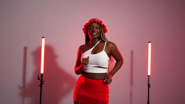 Young energetic african woman with long braids performing modern afrodance moves in a studio with red neon lights, smiling and enjoying the music