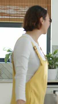 Vertical video: Seeing pal, woman in yellow overalls walking across kitchen counter hugging friend