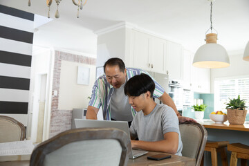 Fototapeta premium Asian father guiding son sitting at kitchen table using laptop and smartphone