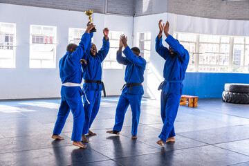 Four african american martial artists in blue gis raising gold trophy, cheering on gym mats