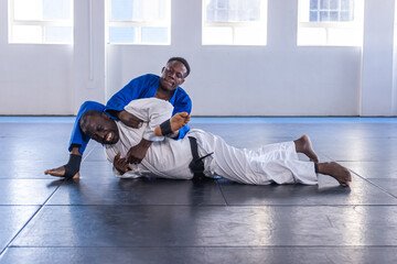 African american male training partners grappling on padded mat in dojo in blue and white gi