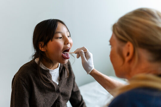 Senior female pediatrician examining girl's mouth with tongue depressor in clinic