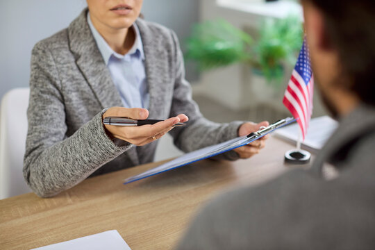 US visa interview at consulate, officer reviewing applicant documents at office desk with United States of America flag. Female lawyer with clipboard and man discuss USA immigration application