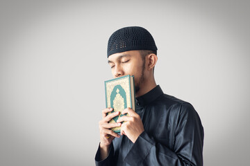 Young Muslim man wearing black thobe, kissing the Quran with respect, isolated on light grey background, symbolizing devotion, faith, and reverence in Islam.