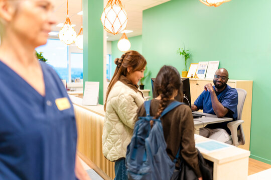 Male receptionist looking at girl standing with mother filling registration form at hospital reception desk