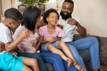 Obraz premium Diverse family sitting on gray sofa in living room, father tickling daughter and mother watching