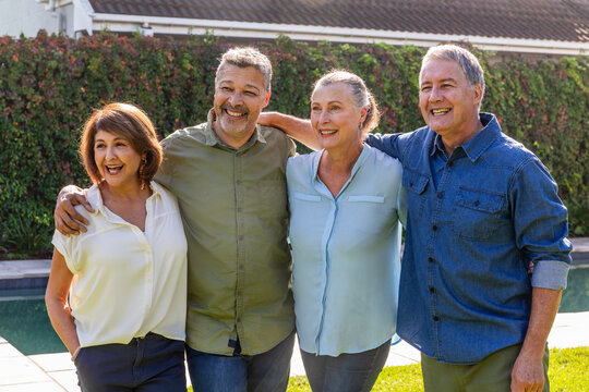 Senior friends hugging shoulders and smiling while standing in backyard by swimming pool and hedge