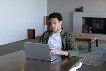 Naklejka premium African american man typing on silver laptop while sitting at wooden table in living area