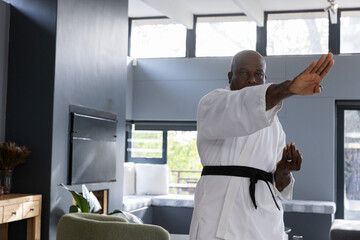Naklejka premium Senior african american man wearing karate gi, belt punching in living room by bench