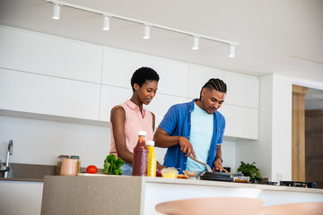 Diverse couple cooking vegetables in frying pan on kitchen island with tomato and cutting board
