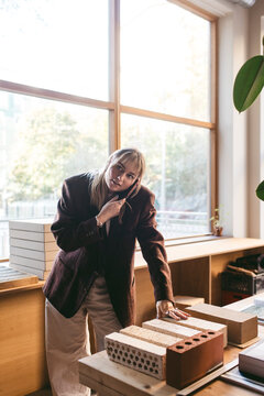 Female design expert talking on smart phone while standing near bricks kept on table in office