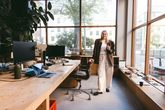 Full length portrait of smiling young businesswoman standing near chair in design company