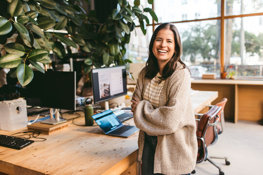 Portrait of happy mature female architect standing with arms crossed near desk in office