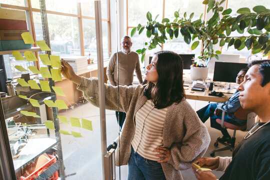 High angle view of mature female design professional explaining ideas from adhesive note on glass door in office meeting