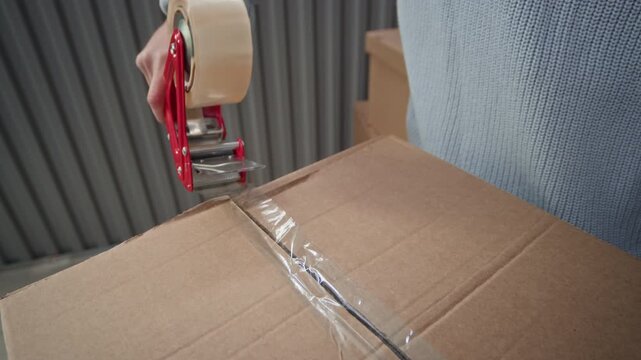Close up view of hands of unrecognizable man sealing cardboard box with red handheld tape dispenser while preparing package for storage or shipping