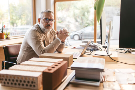 Concentrated mature male design professional sitting with hands clasped at desk in office