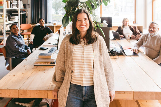 Portrait of smiling female creative professional standing near desk with colleagues sitting in background at office