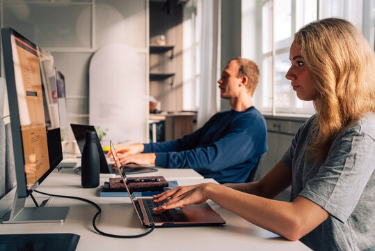 Blond female programmer coding on laptop and computer while sitting at desk in startup office