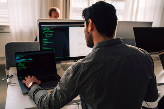 Rear view of young male programmer typing on laptop while sitting at desk in tech office